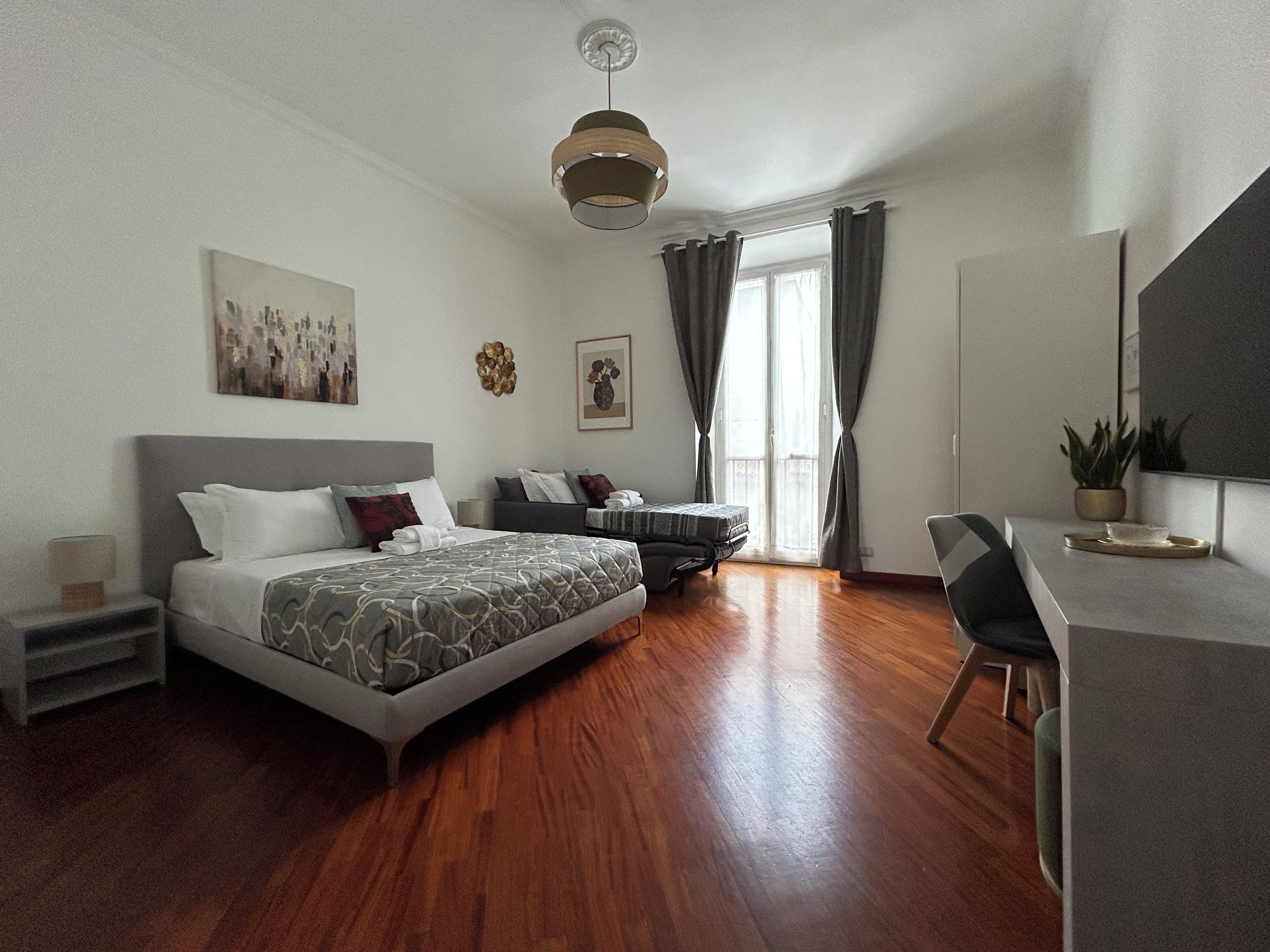 A modern bedroom with a large bed, a desk, and a chair. There is a window with curtains, a ceiling light, and artwork on the wall.