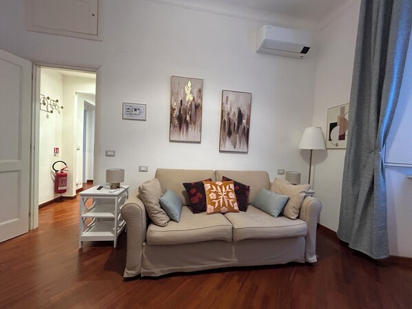 A living room with a beige sofa, two side tables, a fire extinguisher, and abstract wall art. - Domus Vittoria, the ideal apartment for your family in the Vatican Quarter (Roma)