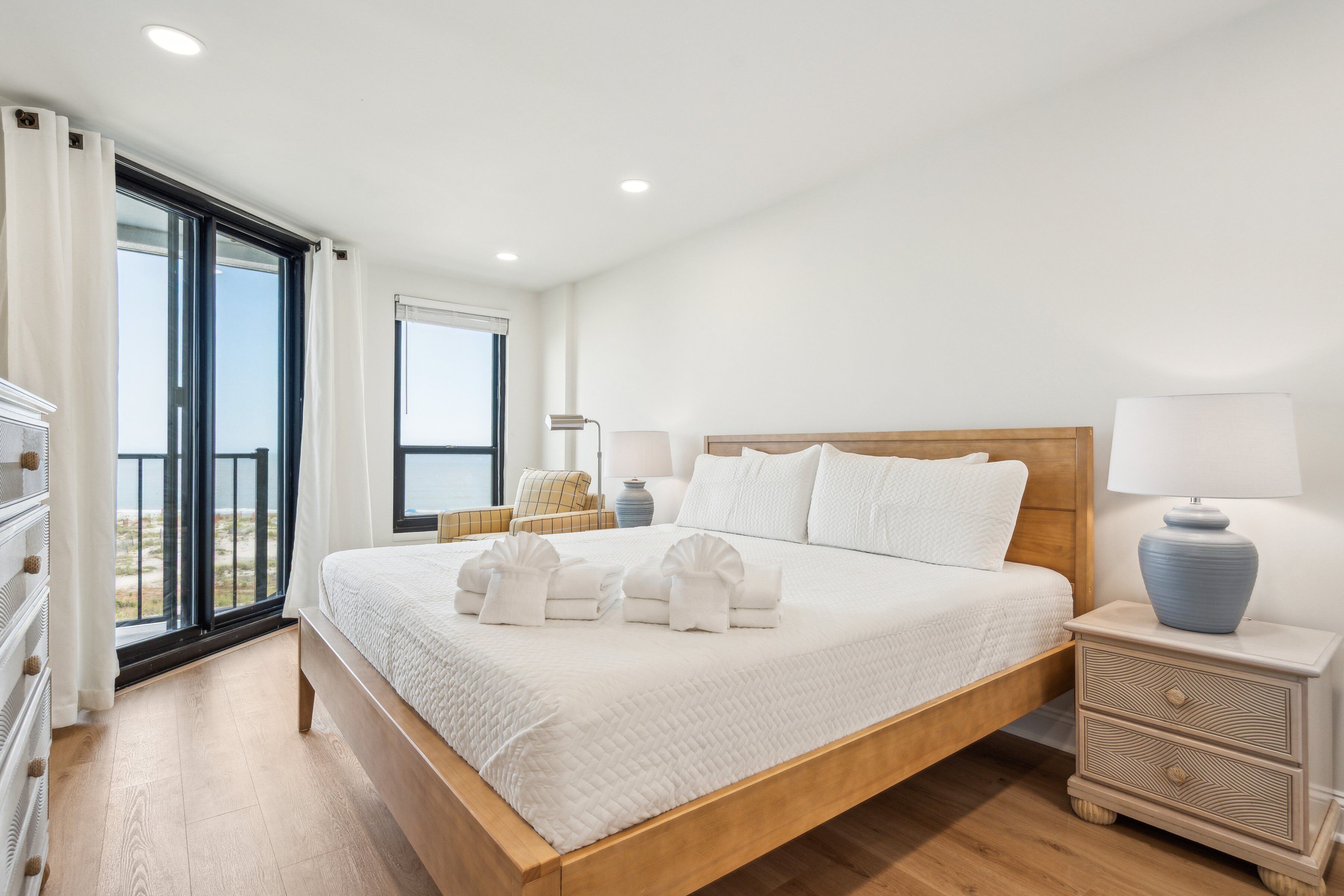 A spacious bedroom with a large bed, a wooden headboard, and a view of the beach through a sliding glass door.