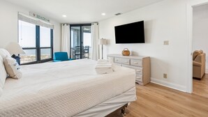 A hotel room with a large bed, a television mounted on the wall, a desk with a lamp, and a view of the beach through the window. - Stunning Ocean Views, Steps from Beach, Modern/Updated Condo- 302 Summerhouse (Isle Of Palms)