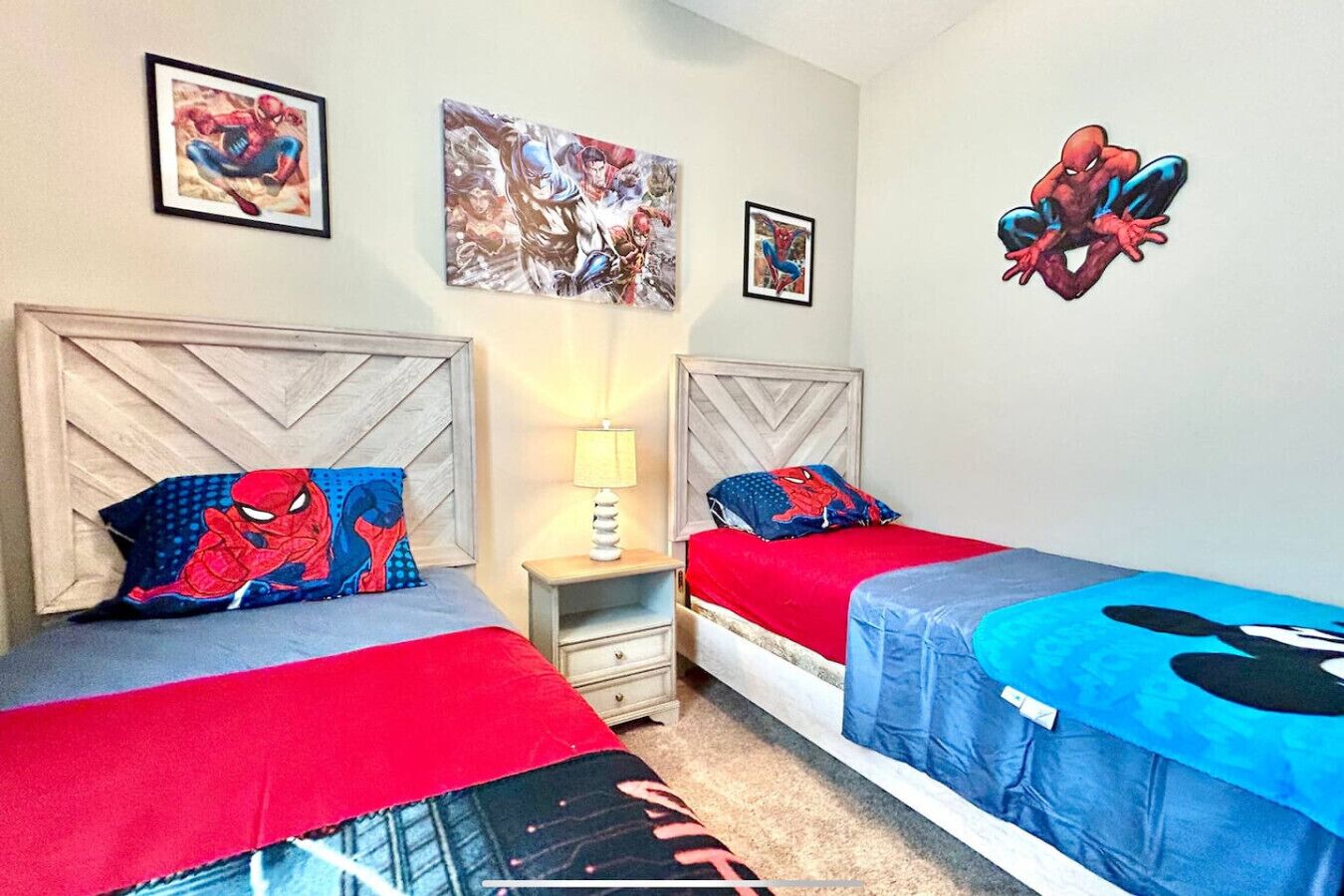 A bedroom with two beds, each with a Spider-Man themed bedspread. There are framed Spider-Man posters on the walls and a lamp on the nightstand.