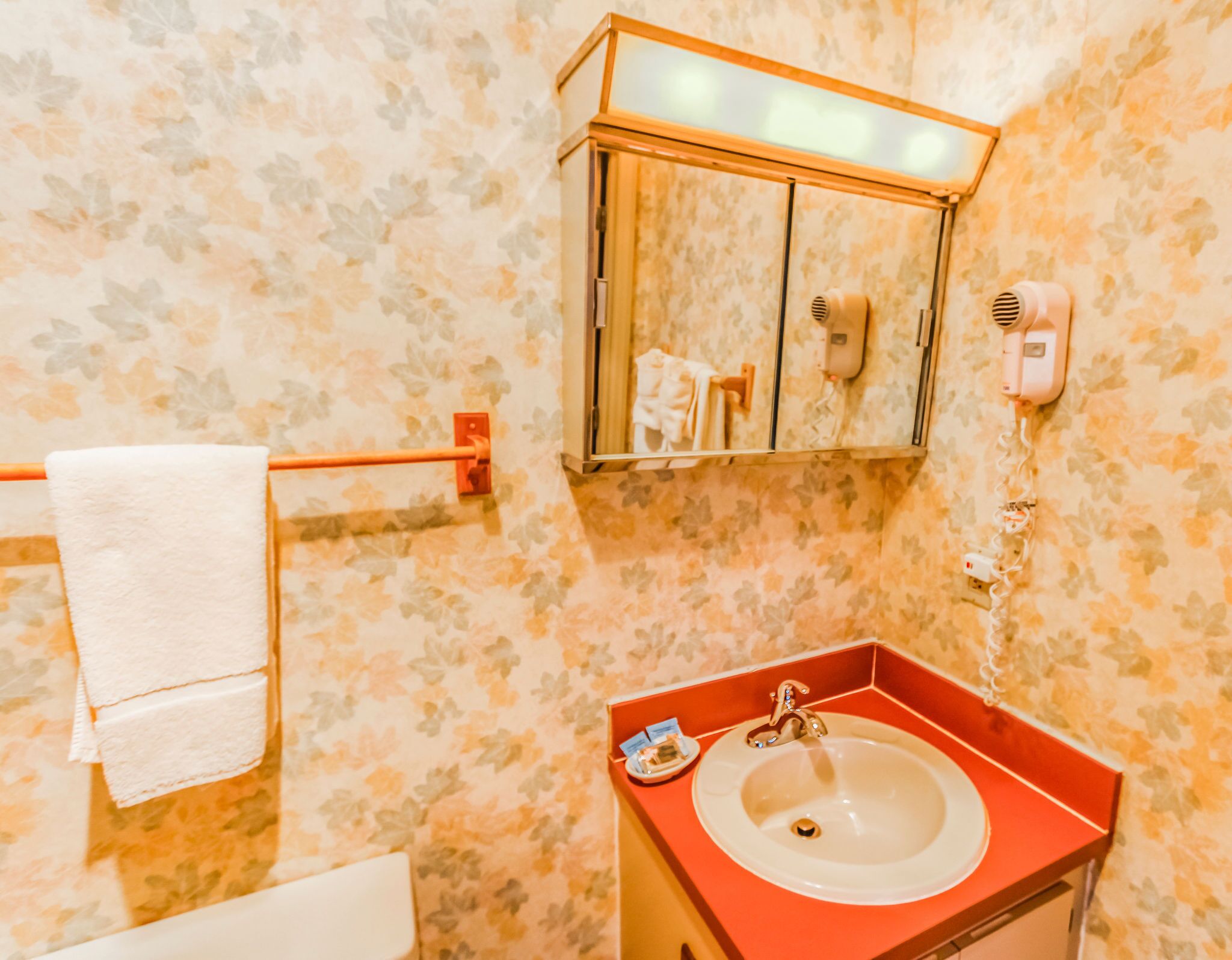A bathroom with a floral wallpaper, a white sink with a red countertop, a towel rack, and a mirror cabinet with a hairdryer and a soap dispenser.