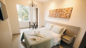 A neatly made bed with a green bedspread, a bedside table, and a nightstand. There is a window with a view of trees and a chandelier. - JARDIM DO RIO SUÍTES (Florianopolis)