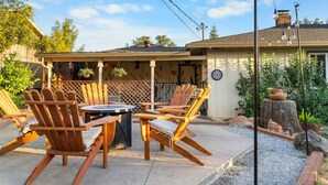 Un patio al aire libre con sillas de madera y una mesa redonda, rodeado por una valla de madera y macetas con plantas.