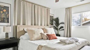 A bedroom with a bed, bedside tables, a nightstand, a window, and a plant.