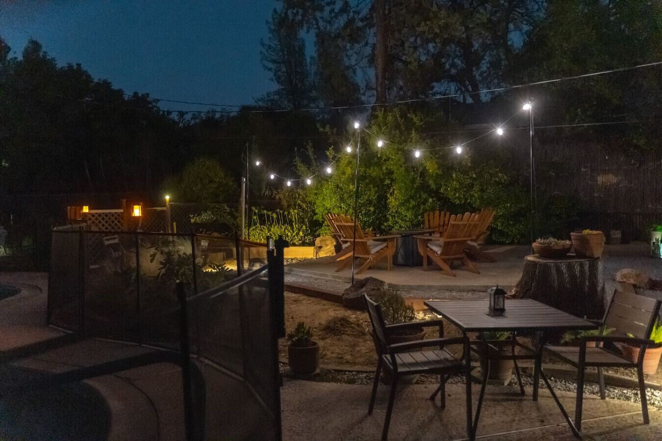 En udendørs patio med lyskæder, træstole, et bord og plantekasser.