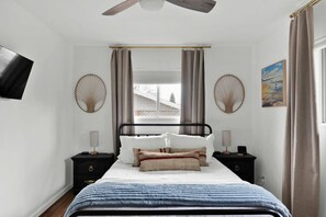 Quarto com cama, duas mesas de cabeceira, ventilador de teto, televisão e janela com cortinas.