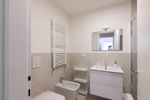 Une salle de bain comprenant des toilettes, un bidet, un lavabo, un miroir, un porte-serviettes et un espace douche.