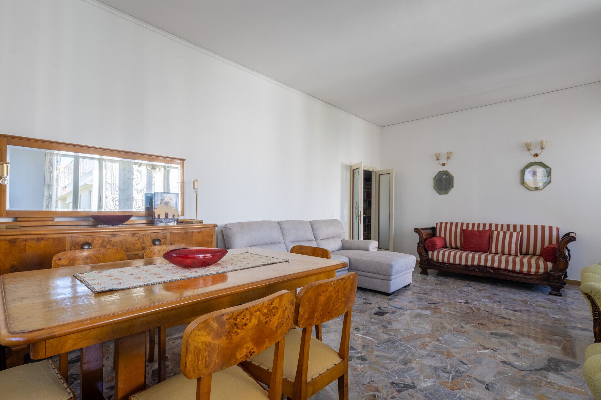 A living room with a wooden dining table, two chairs, a grey sofa, a red and white striped sofa, and a mirror on the wall.