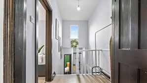 Un pasillo con piso de madera, barandal blanco y una puerta que da a una habitación con cama y pared verde.
