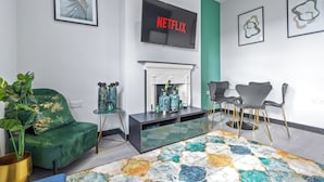 Una sala de estar moderna con un sofá verde, una chimenea, una mesa de centro de vidrio y un televisor de pantalla plana mostrando 'NETFLIX'.