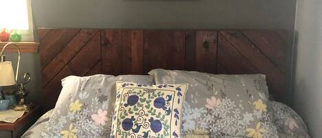 Una cama bien tendida con almohadas estampadas con motivos florales, un cuadro colgado en la pared que muestra un puente, y un cabecero de madera.