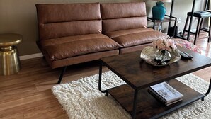 Ein Wohnzimmer mit braunem Ledersofa, einem Couchtisch aus Holz mit dekorativen Gegenständen, einem weißen Shaggy-Teppich und zwei abstrakten Bildern an der Wand.