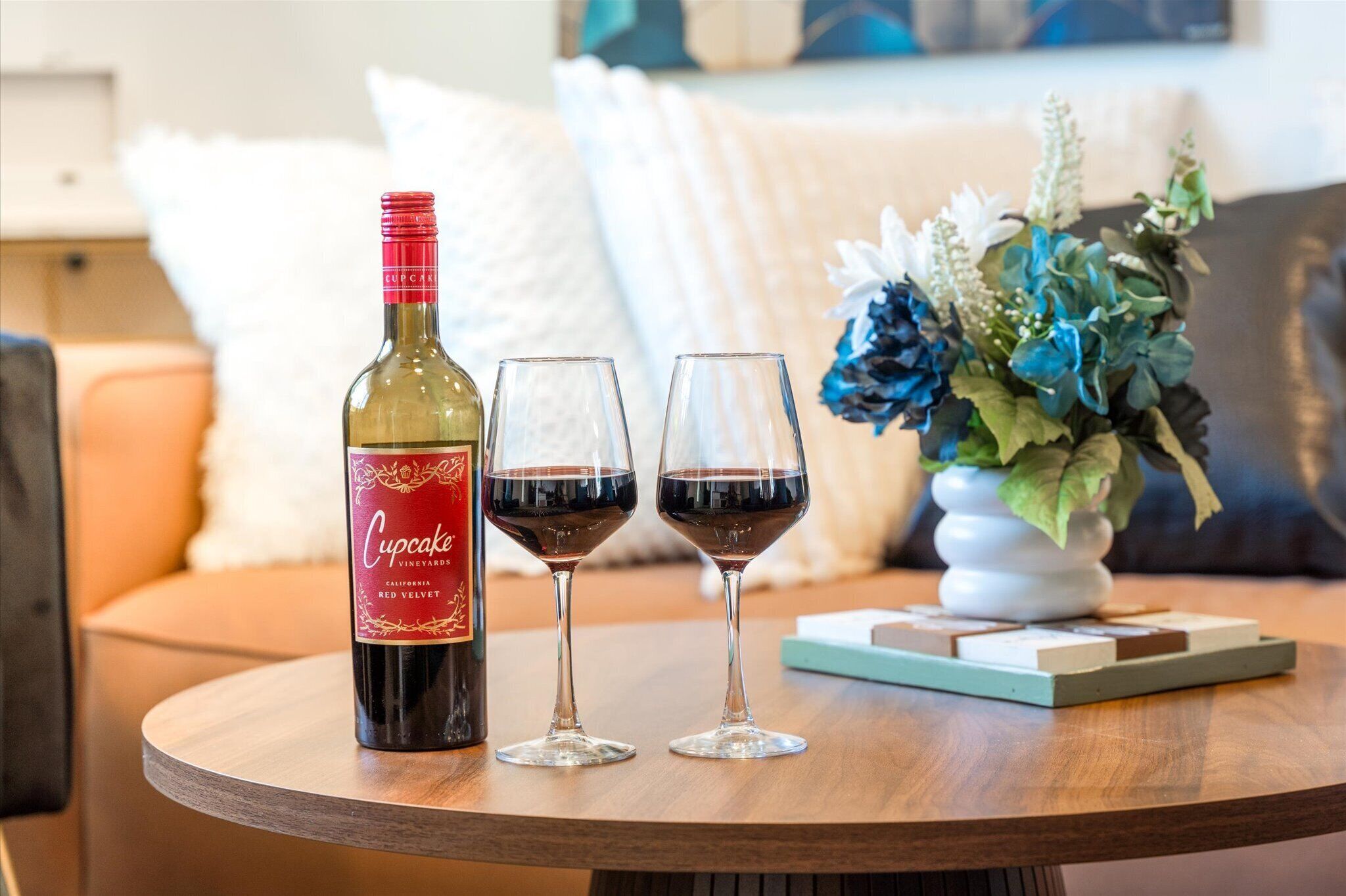Une bouteille de vin rouge «Red Velvet» de la marque Cupcake Vineyards, deux verres à vin et un vase décoratif avec des fleurs, le tout posé sur une table ronde en bois.