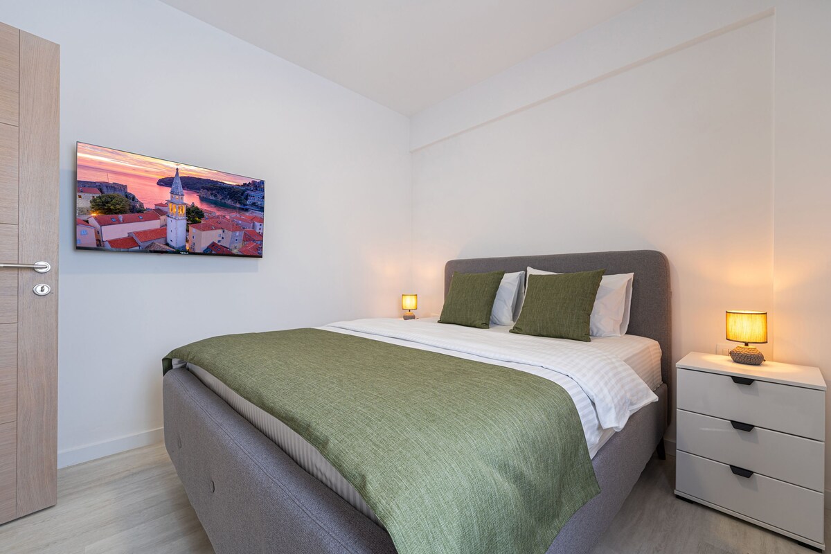 A bedroom with a bed, bedside tables, a nightstand, and a painting of a coastal town.