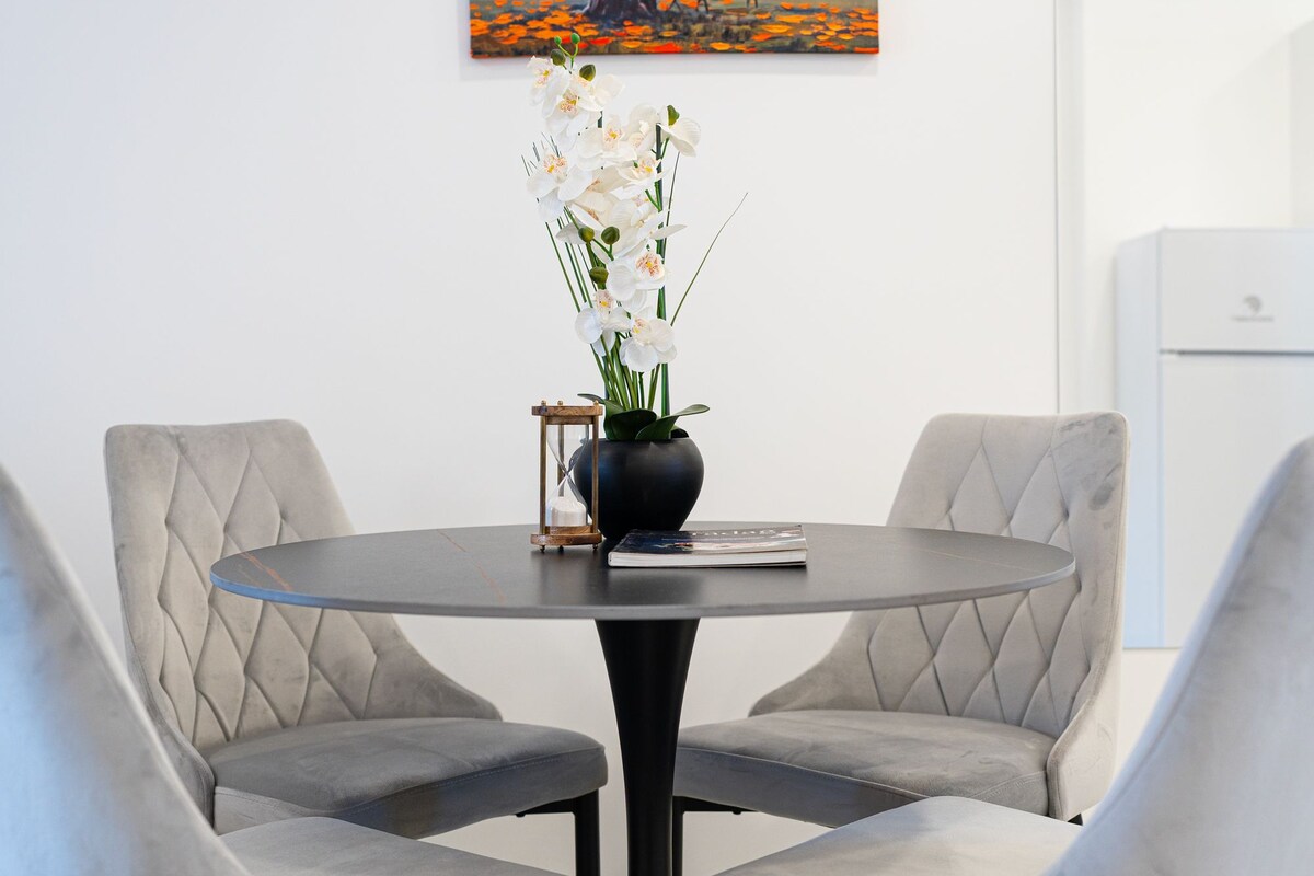 A dining area with a round table, two upholstered chairs, a vase with flowers, and a painting on the wall.
