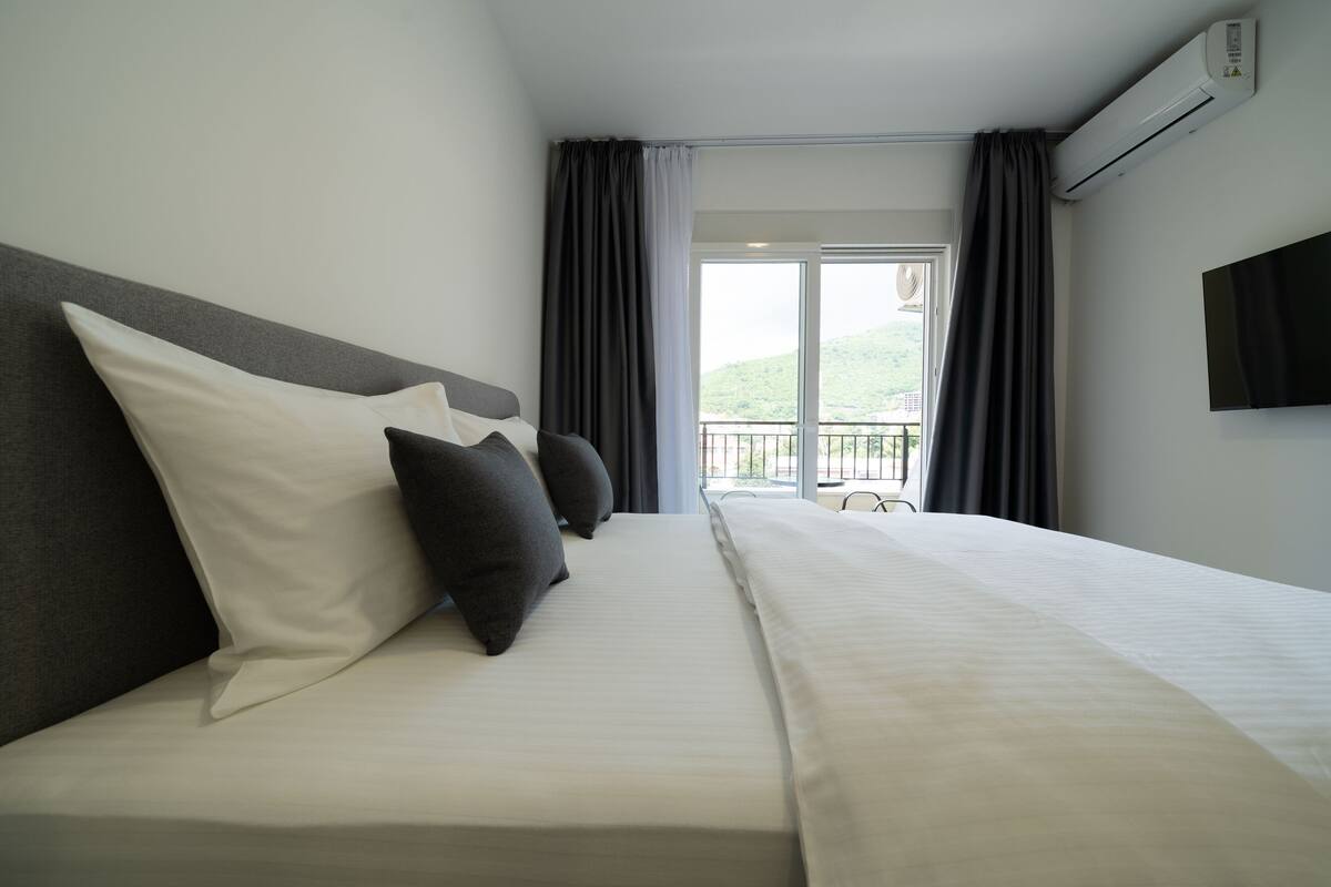 A hotel room with a large bed, a flat-screen TV, and a view of a balcony with a railing.