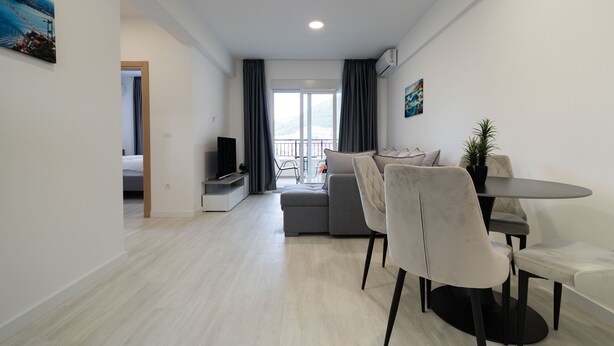 Ein modernes Wohnzimmer mit grauem Sofa, einem Esstisch mit Stühlen, einem Fernseher und einem Balkon mit Vorhängen.
