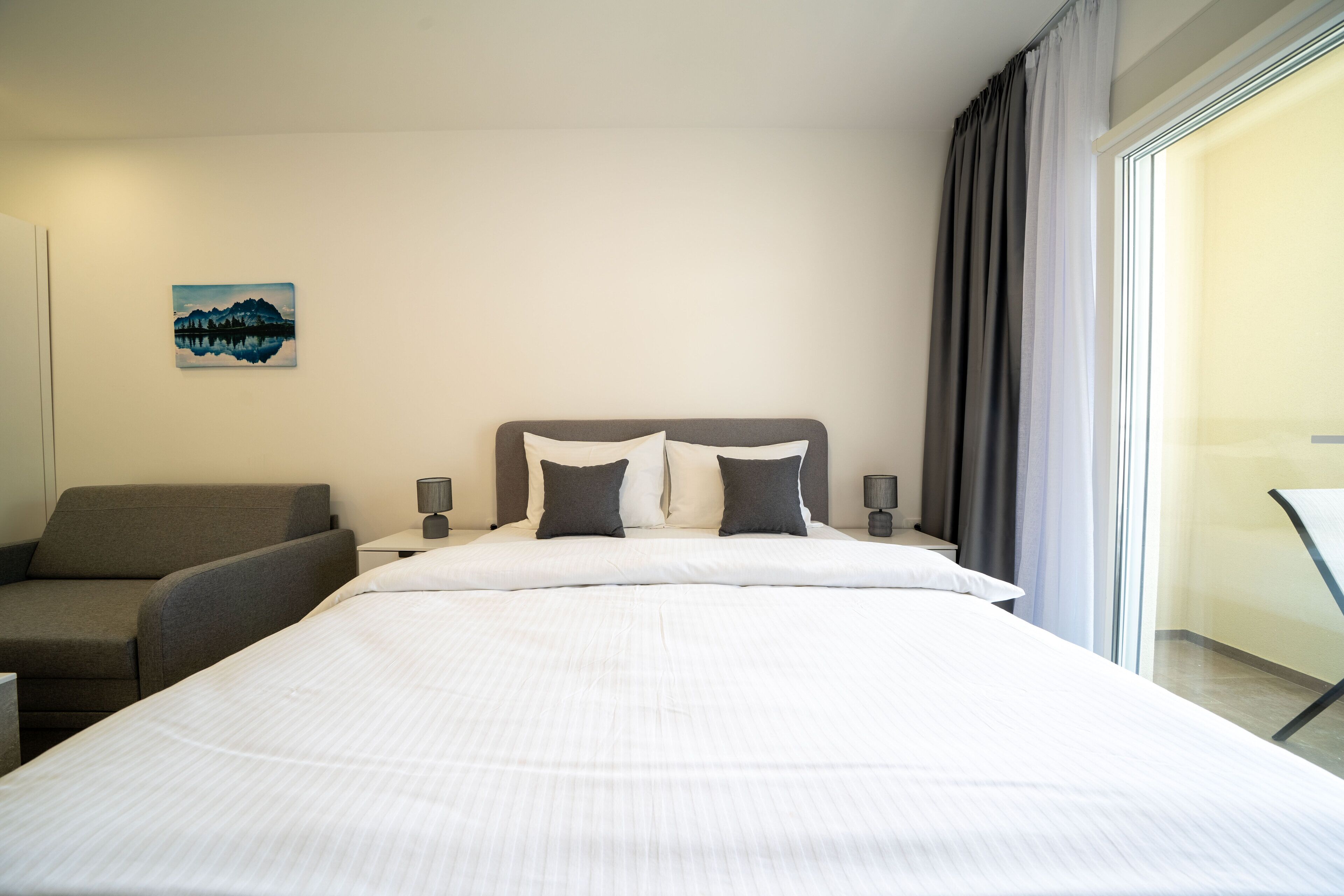 Ein Hotelzimmer mit einem großen Bett, zwei Nachttischen mit Lampen, einem grauen Sessel und einem Bild an der Wand.