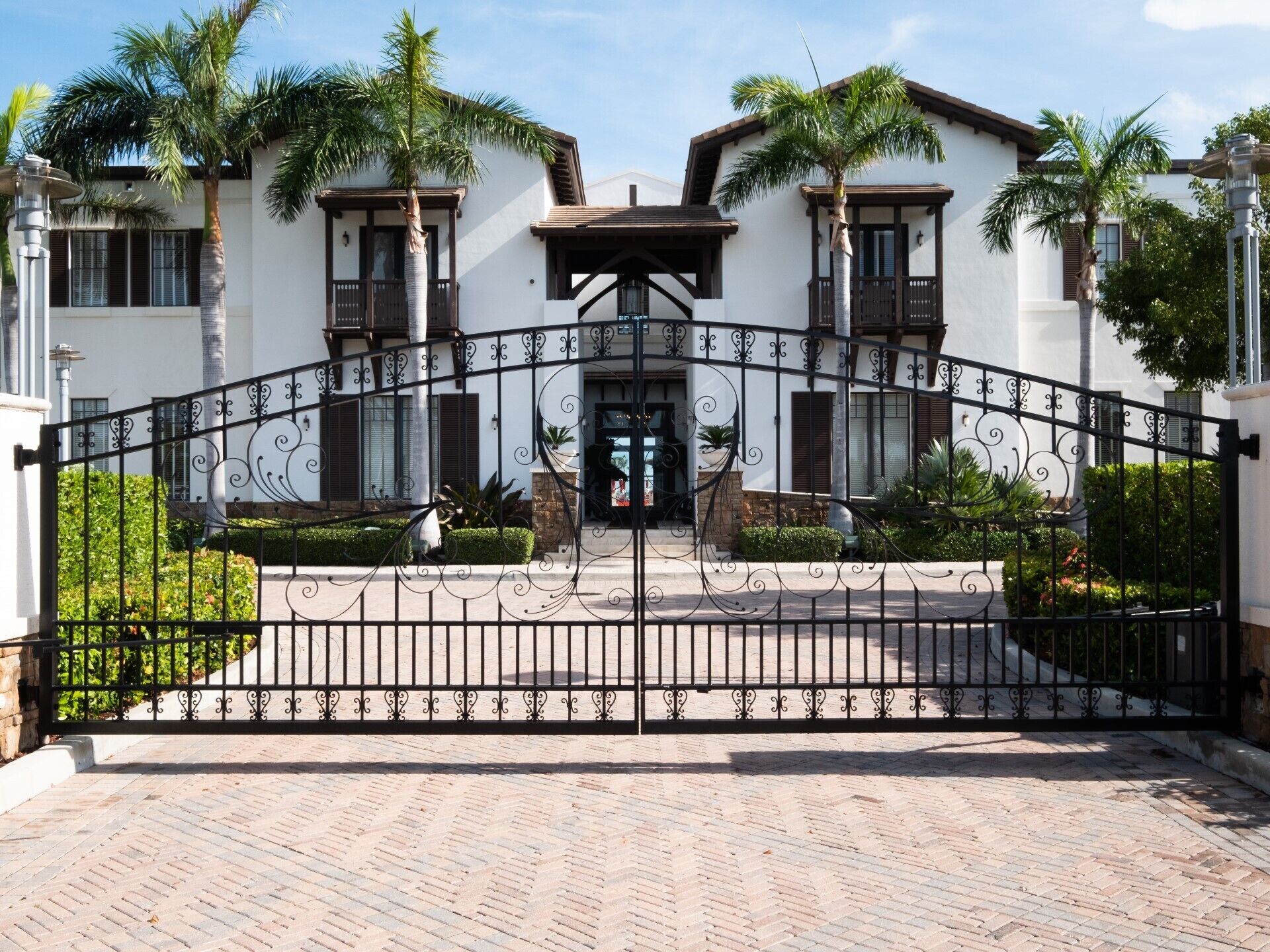 A gated entrance with a wrought iron gate, palm trees, and a building with a white facade and brown trim.