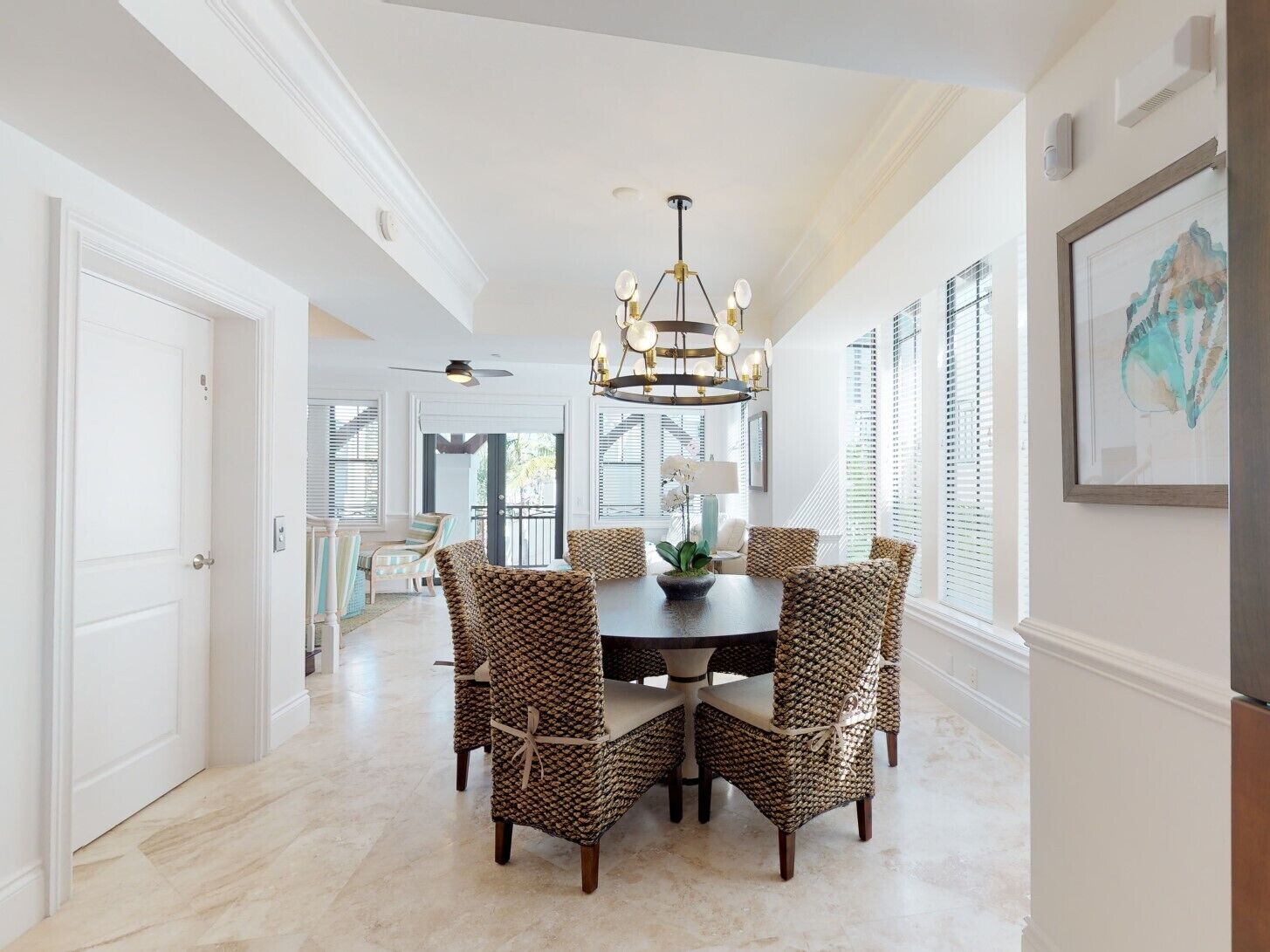 A dining area with a round table, six wicker chairs, a chandelier, and a large window with blinds.
