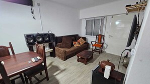 A living room with a sofa, a coffee table, a dining table with chairs, a speaker system, and a television mounted on the wall.