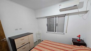 A bedroom with a bed, a dresser, a chair, a window with blinds, and an air conditioning unit.