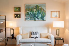 Uma sala de estar com um sofá, duas mesas laterais com luminárias, um quadro na parede e uma estante de livros.