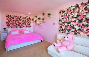 Un dormitorio con cama, sofá blanco, oso de peluche rosa y decoración floral en la pared.