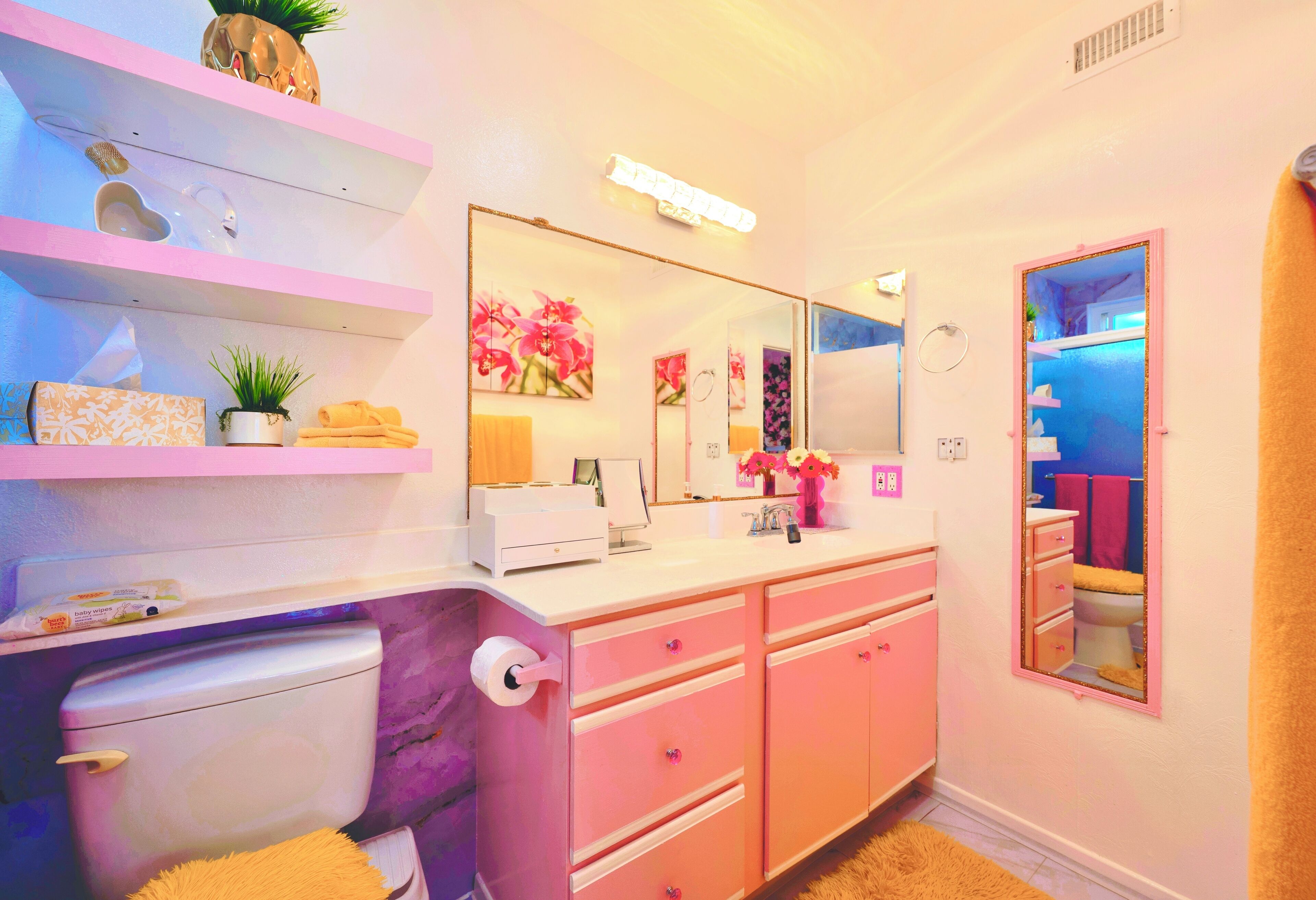 A bathroom with a toilet, sink, and mirror. There are pink cabinets and a shelf with toiletries.