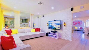 Un salon moderne avec un canapé blanc, des coussins rouges et jaunes, un téléviseur à écran plat et un panneau décoratif mural.