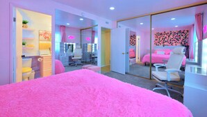 Une chambre décorée dans des tons de rose, avec un lit, un bureau et une chaise. On y trouve un miroir, une porte et un mur orné d’un motif floral.
