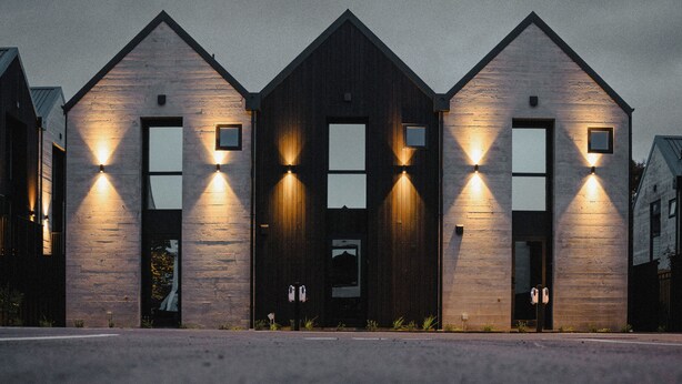 Una hilera de tres edificios modernos con fachada oscura y ventanas iluminadas.
