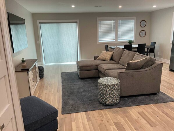 A living room with a sectional sofa, a round ottoman, and a dining area with a table and chairs. - The Landing@Clinton River (Harrison Twp)