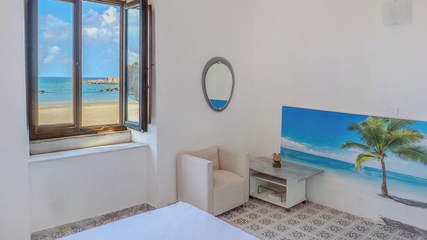 Ein Zimmer mit großem Fenster mit Blick auf einen Strand, ein kleiner weißer Stuhl, ein Nachttisch und ein Wandbild einer Strandlandschaft.