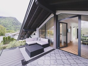 Un área de estar exterior moderna con un sofá y cojines, una terraza de madera y un ventanal que da a un espacio interior.