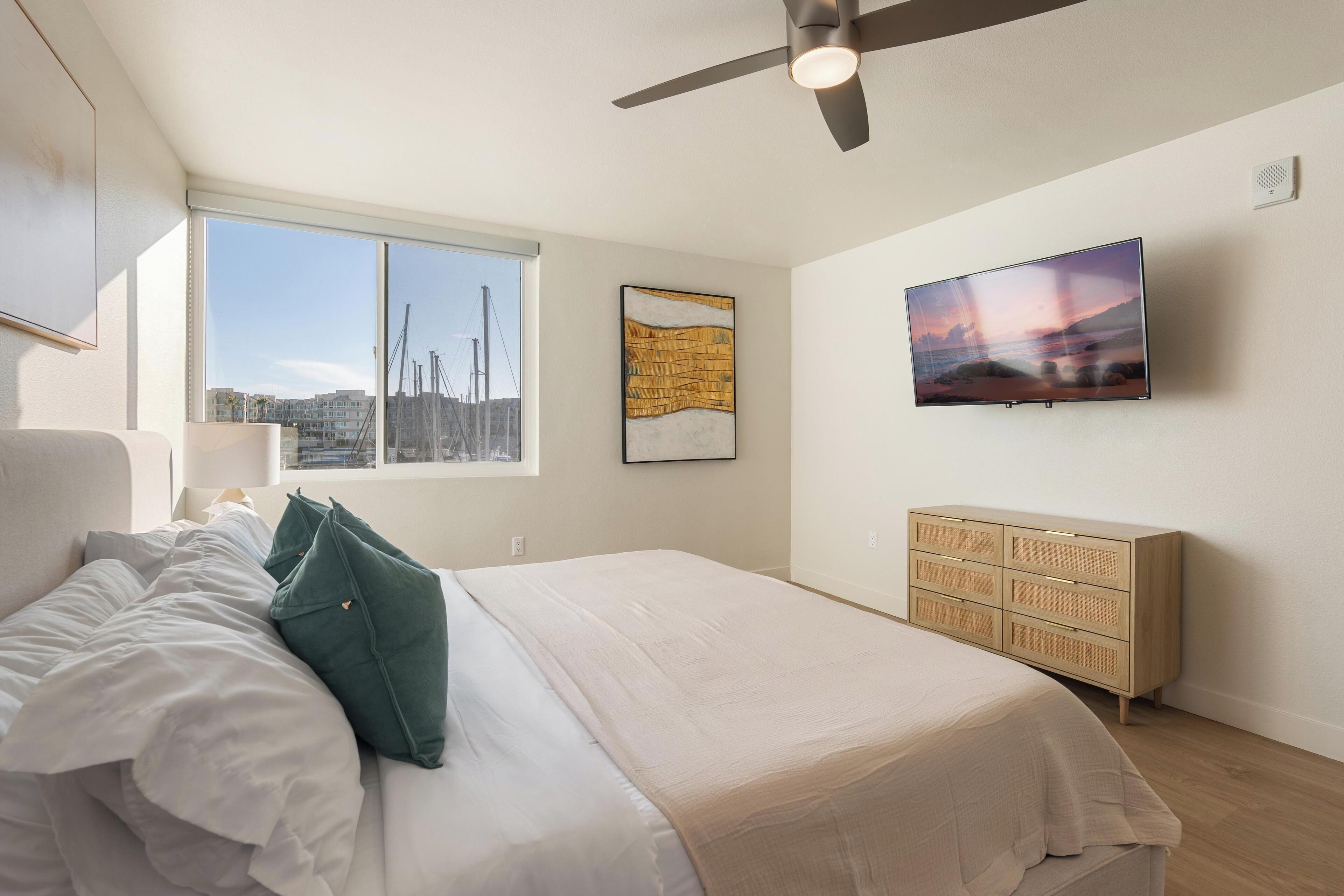 Ein Schlafzimmer mit einem Bett, einem Deckenventilator, einem Fernseher, einem Fenster mit Stadtblick und einer Holzkommode.