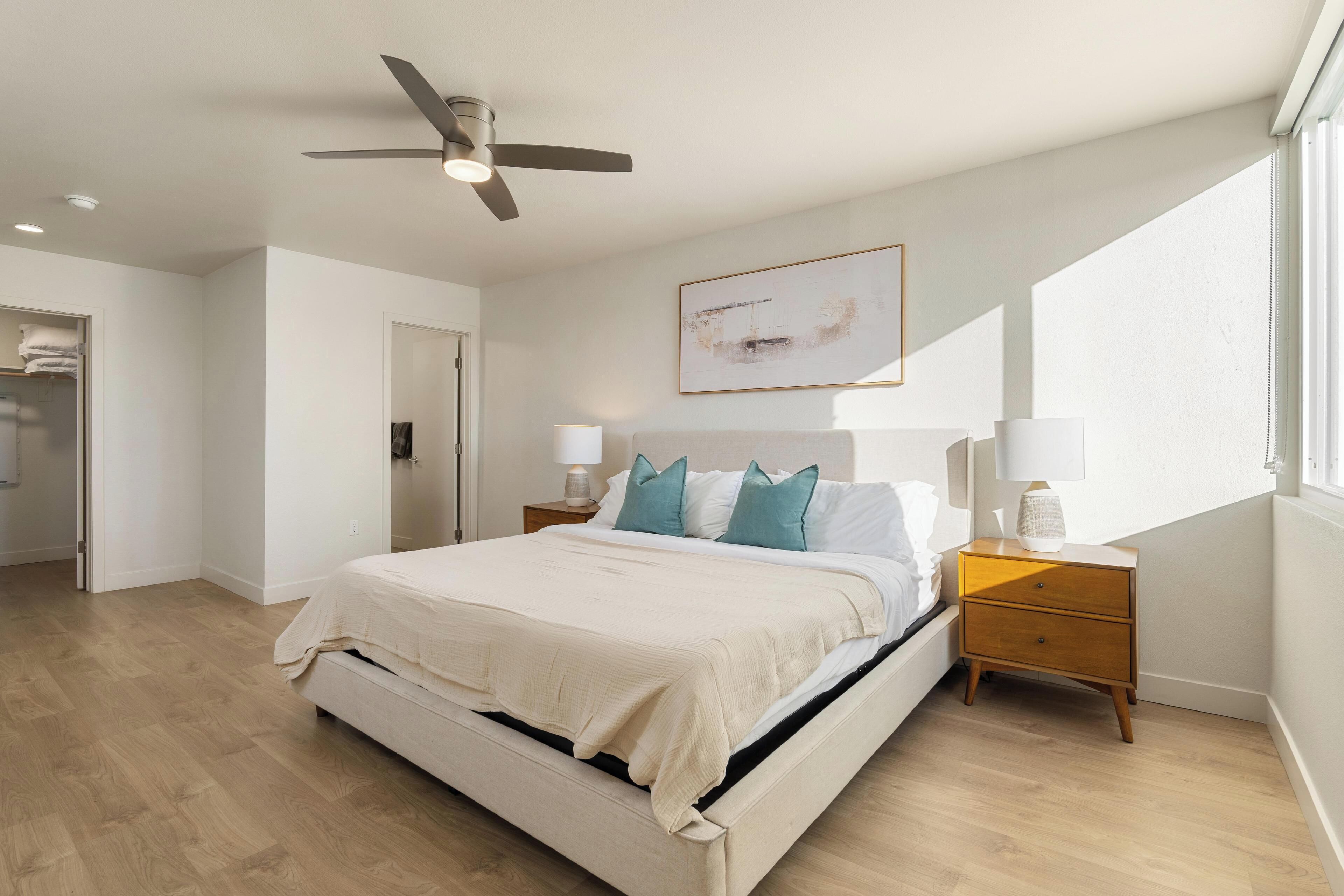 Un dormitorio con cama, mesitas de noche, ventilador de techo, un cuadro y una ventana.