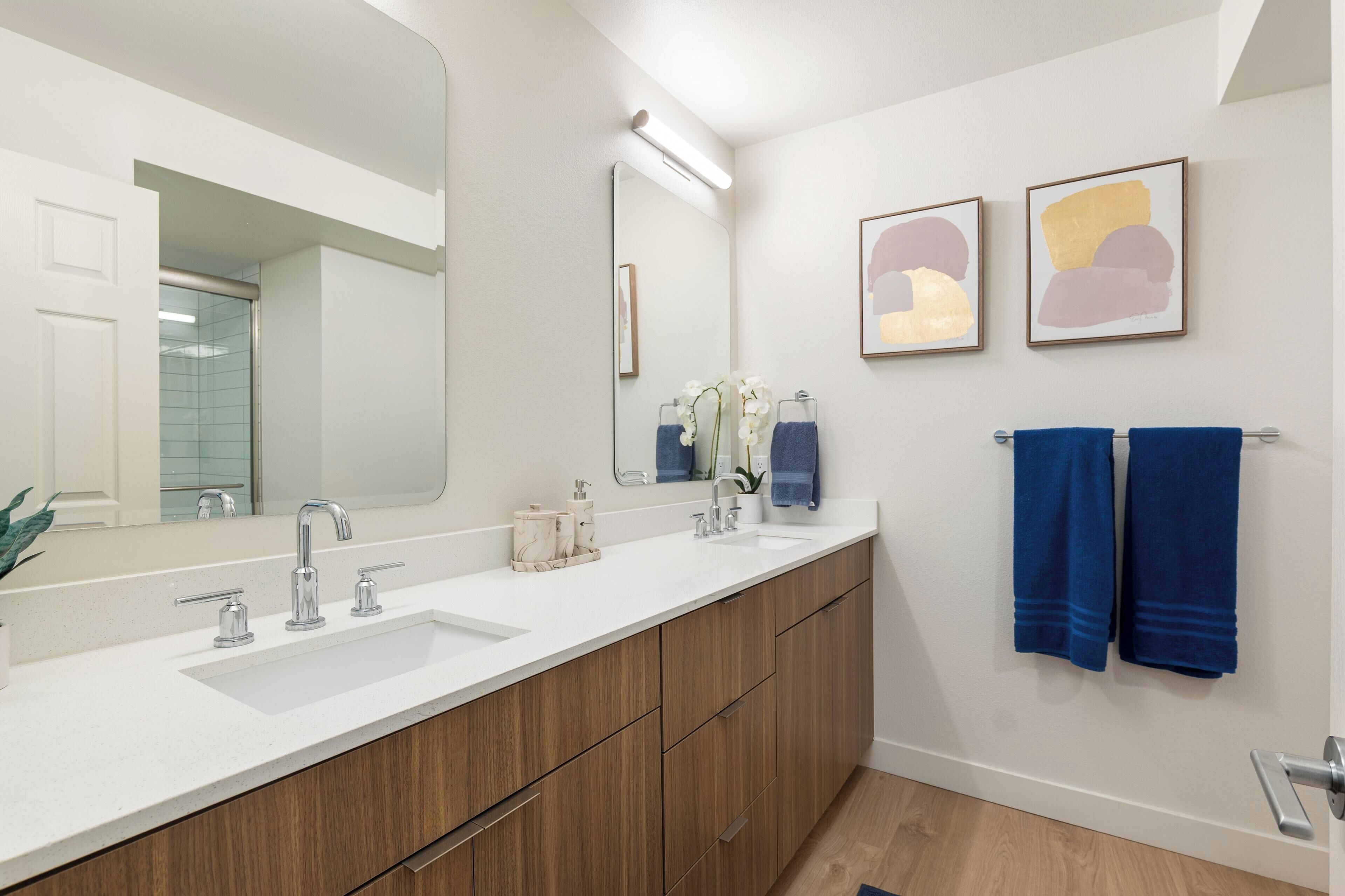 Un baño moderno con lavamanos doble, armarios de madera, un espejo grande y dos toallas colgadas en un perchero.