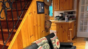 Un salon chaleureux avec un canapé en cuir noir, des murs en bois et une cuisine à l’arrière-plan.
