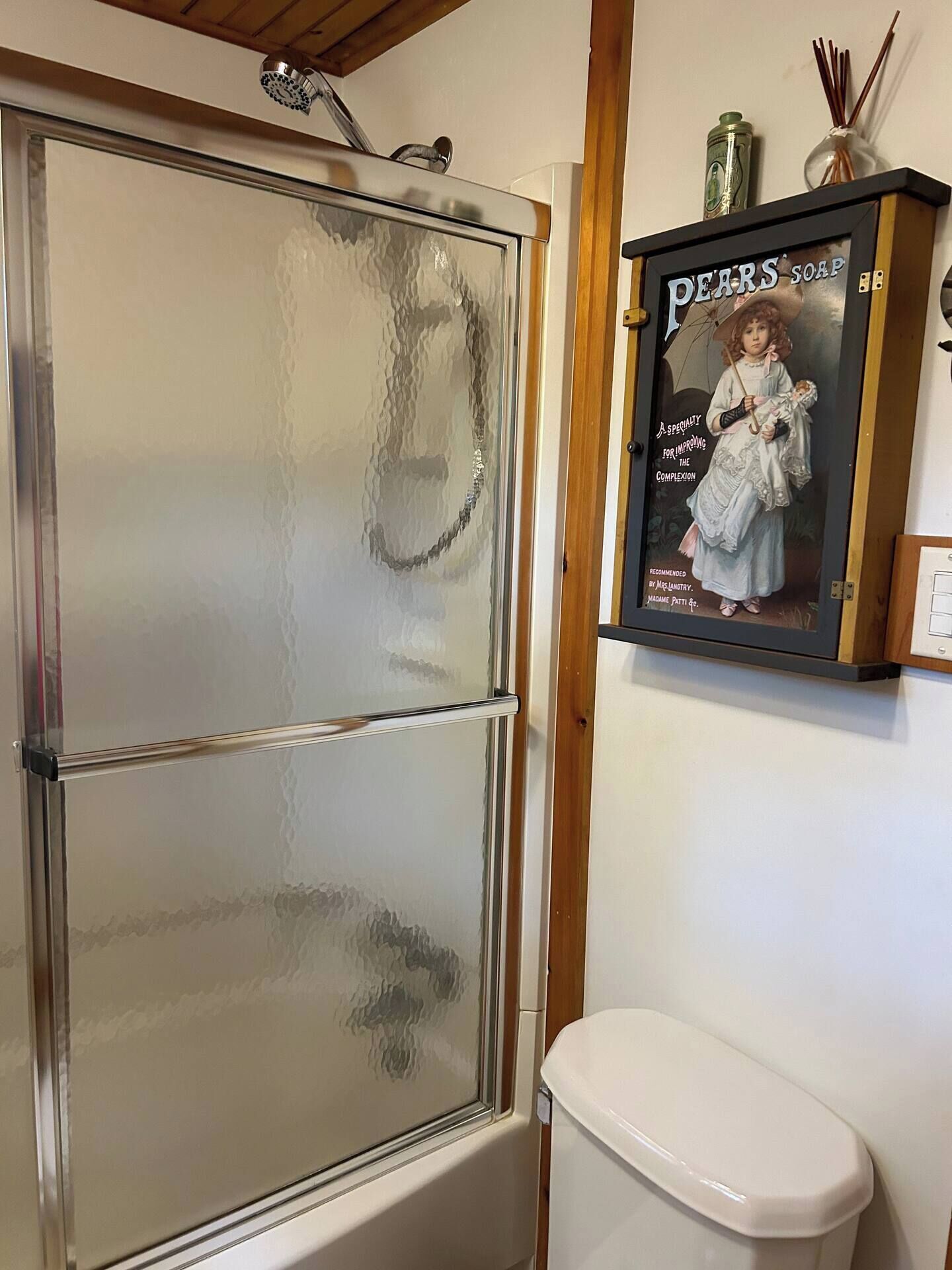 Un baño con cabina de ducha de vidrio, un inodoro blanco y un anuncio enmarcado de Jabón Pears.
