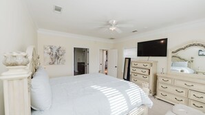 Een slaapkamer met een groot bed, een plafondventilator, een dressoir, een televisie en een deur naar een andere kamer.