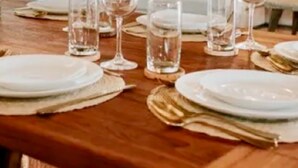Una mesa de comedor preparada con platos, vasos y cubiertos, con una planta y un sofá al fondo.