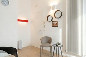 Une chambre d’hôtel moderne avec un plancher en bois, un fauteuil gris, une petite table ronde avec une plante, une bande lumineuse rouge, une horloge murale, deux miroirs ronds et un tableau encadré.