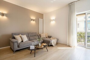 A modern living room with a grey sofa, a coffee table with a vase and snacks, and a view of a balcony through a large window. -  Central Station Apartment – Modern Comfort in the Heart of Bari (Bari)