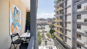 Un balcon avec une table et des chaises, donnant sur un paysage urbain avec des immeubles et une place.