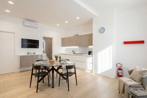A modern kitchen with white cabinets, a dining area with a round table and chairs, a flat-screen TV, and a red fire extinguisher. -  Central Station Apartment – Modern Comfort in the Heart of Bari (Bari)