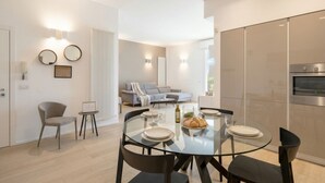 Une cuisine moderne dotée d’un four encastré, un coin repas avec une table et des chaises en verre, et un salon avec un canapé et une table basse.