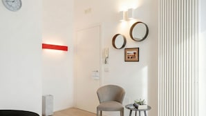 A modern hotel room with a wooden floor, a grey armchair, a small round table with a plant, a red light strip, a wall clock, two round mirrors, and a framed picture. - Central Station Apartment – Modern Comfort in the Heart of Bari (Bari)