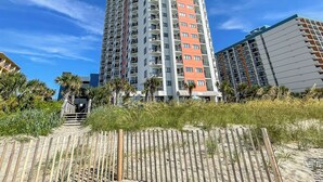 A tall, multi-story building with balconies, located near a sandy area with a wooden fence. - Sunset Palace Perfect Beach Getaway: Stunning Updates & Unbeatable Convenience! (Myrtle Beach)